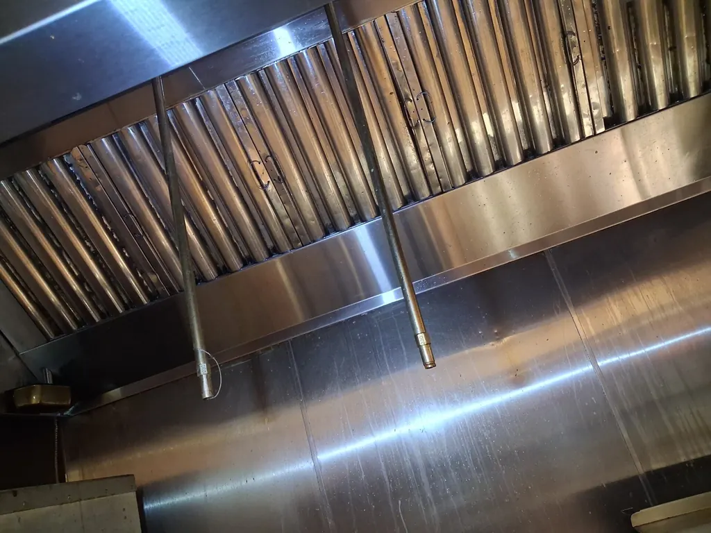 Baffle filters seated in a commercial kitchen hood, still carrying a brown film of grease, with overhead pipework crossing in front of them.