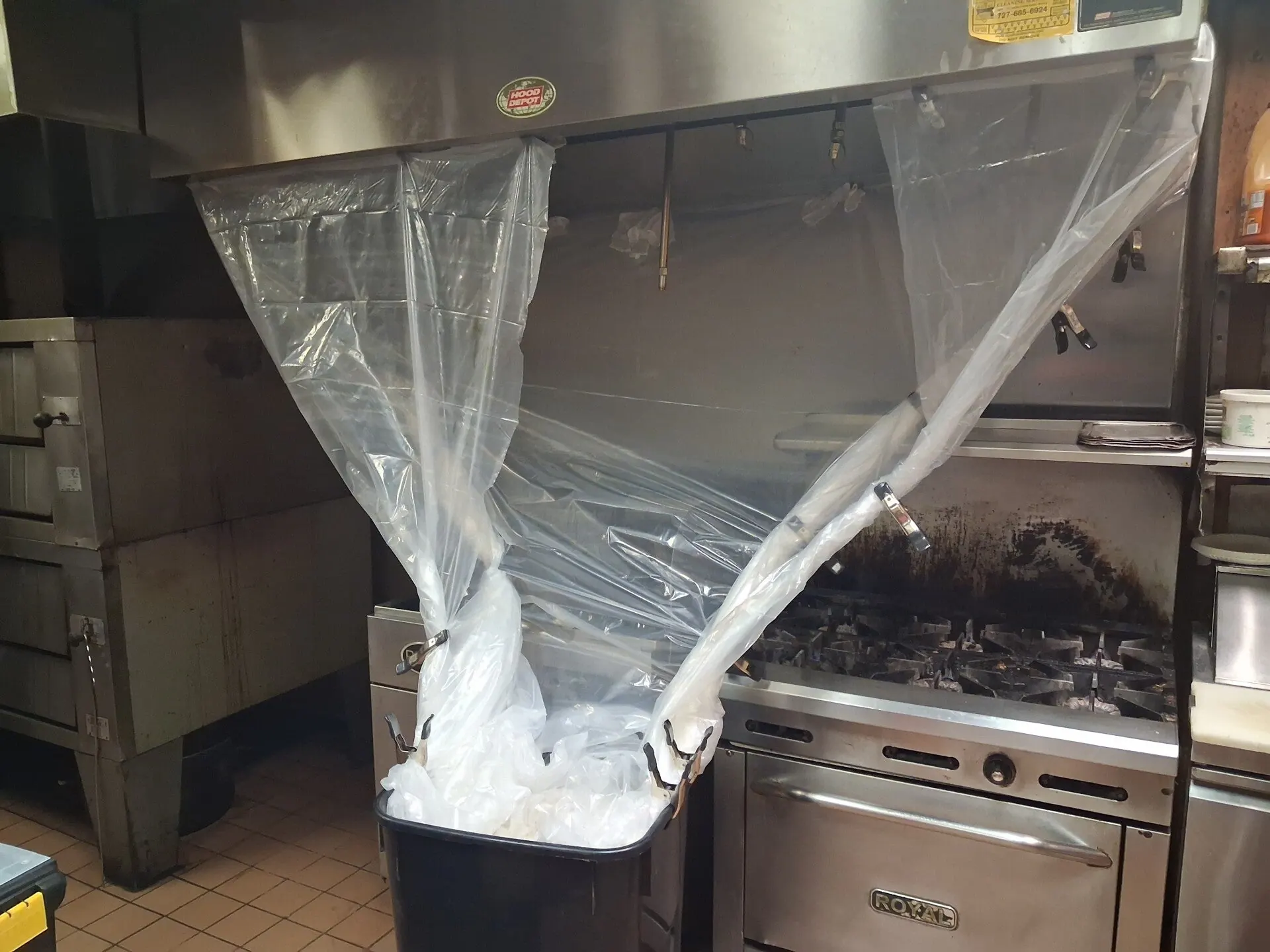 A commercial kitchen extraction hood masked off in clear plastic sheeting that funnels down into a bin, with a greasy range and ovens either side.